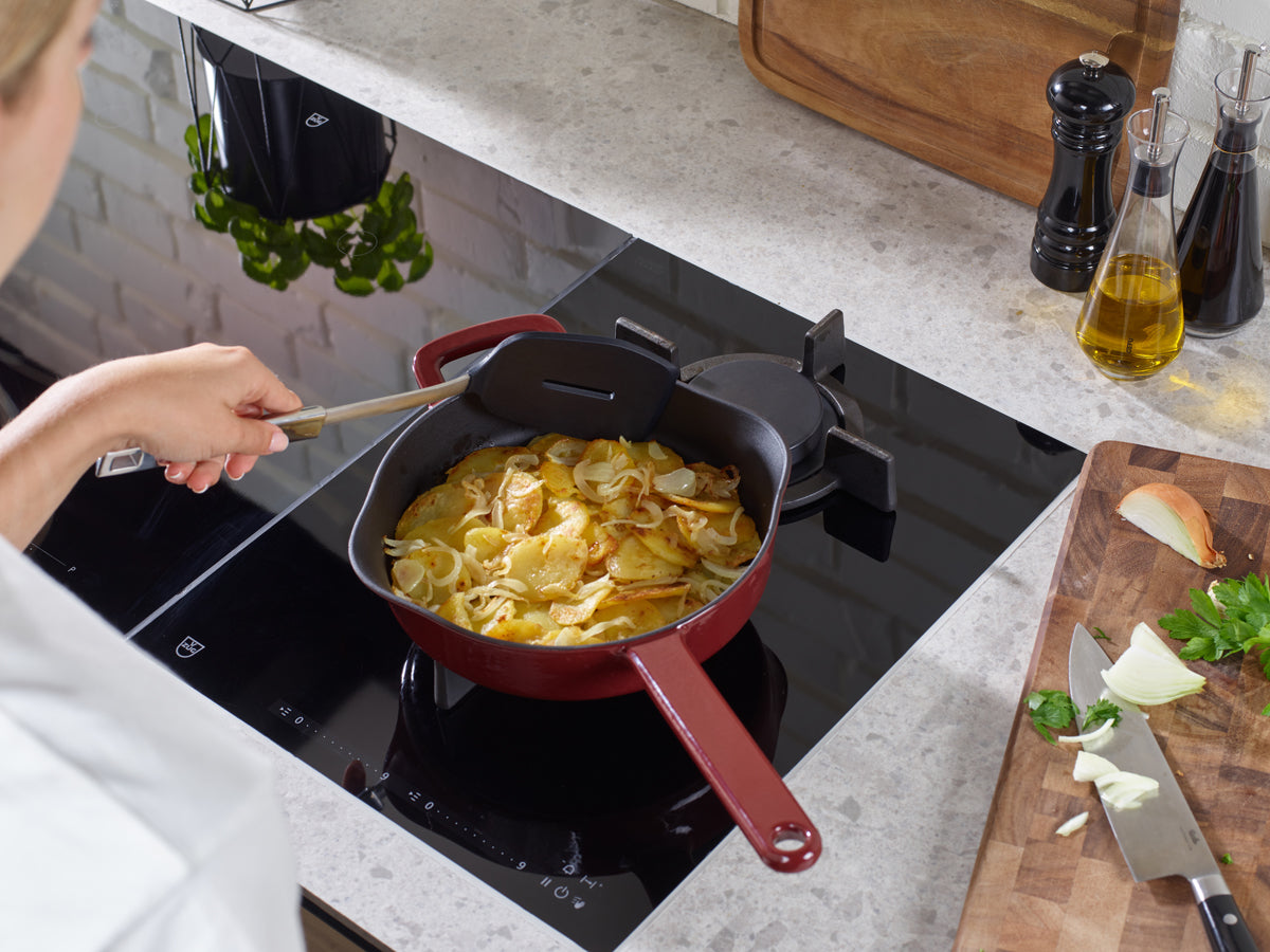 A person stirs potatoes and onions in a red Fissler cast-iron Frying Pan; fresh ingredients and shakers complete this modern kitchen scene.