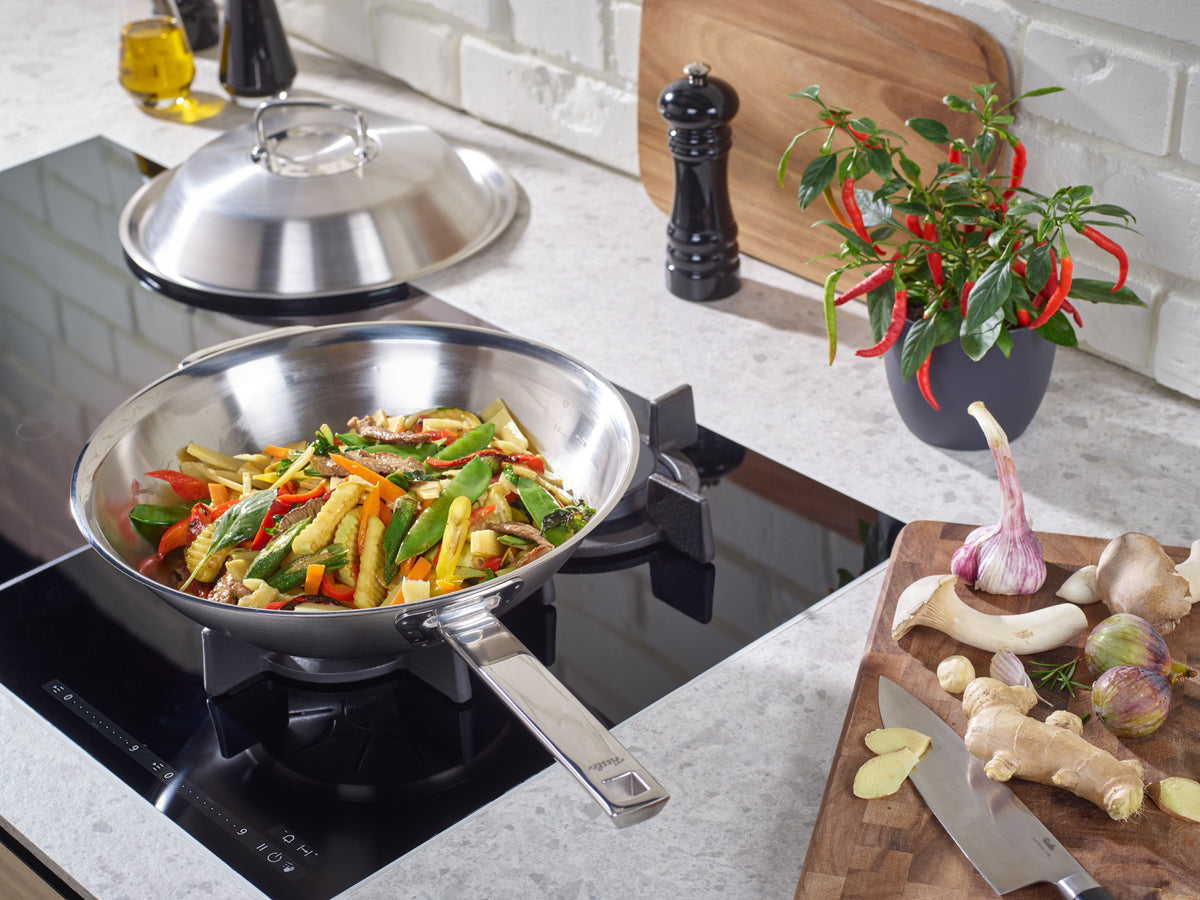 Fissler Original-Profi Collection® Wok 30cm mit Deckel auf dem Herd, Gemüse anbraten; hochwertige Edelstahlpfannen und Gewürze in der Nähe.