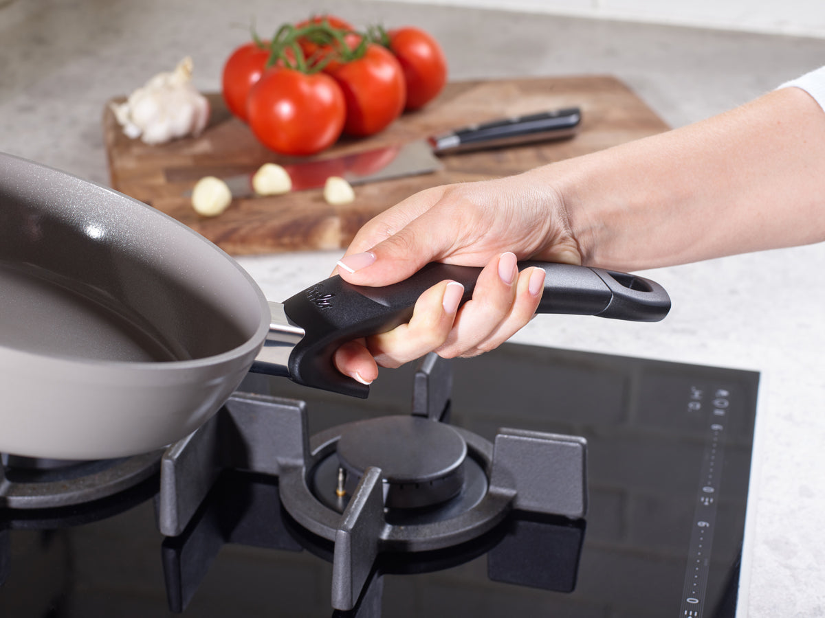 Eine Hand befestigt einen Griff an den Fissler Ceratal® Comfort Pfannen auf dem Herd; frische Zutaten und ein Messer liegen auf einem Holzbrett daneben.