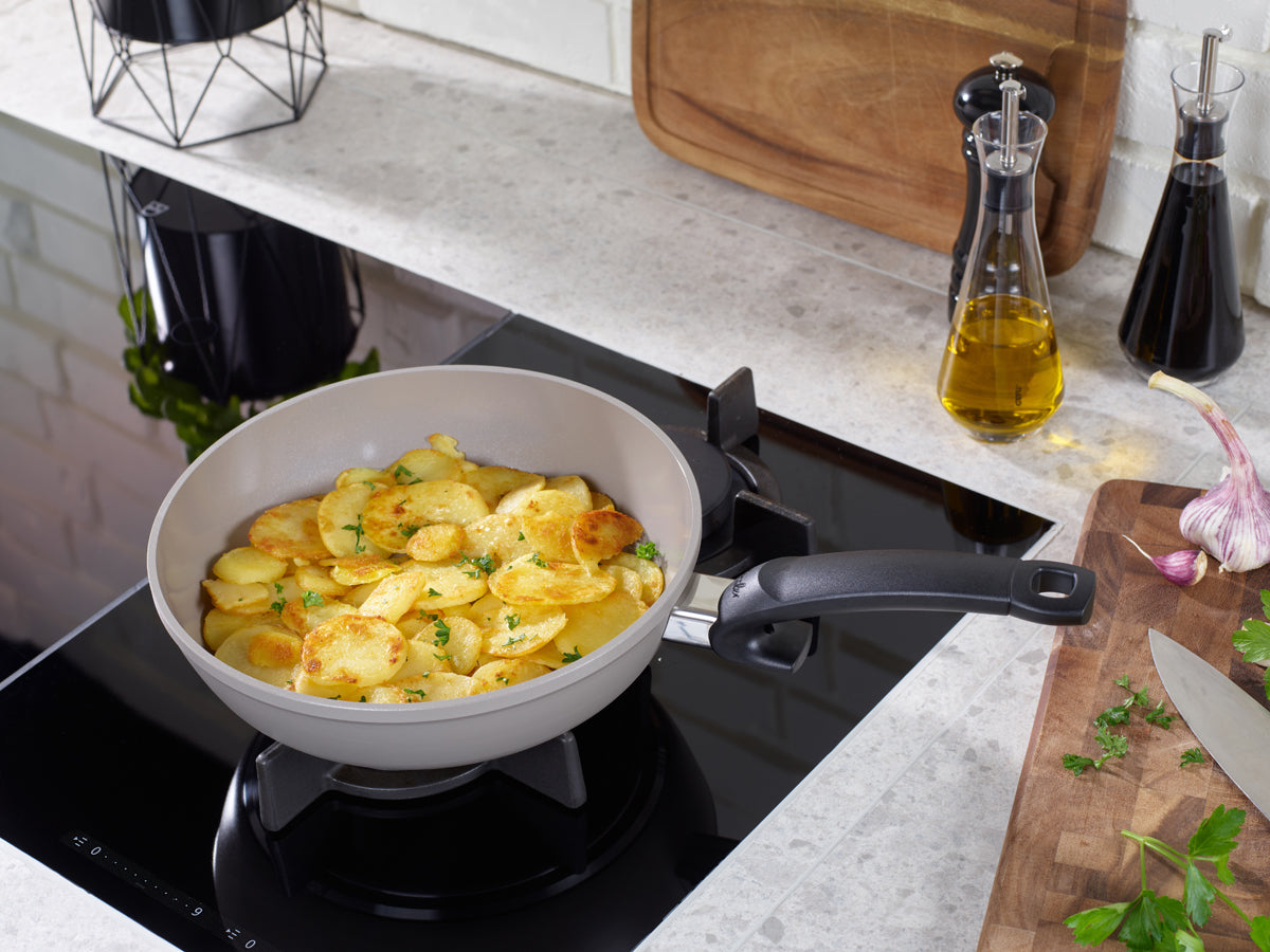 Fissler Ceratal® 2-tlg. Pfannenset kocht Kartoffeln mit Kräutern, deutsches Qualitätsdesign, mit Öl, Knoblauch, Petersilie auf einer hellen Arbeitsfläche.