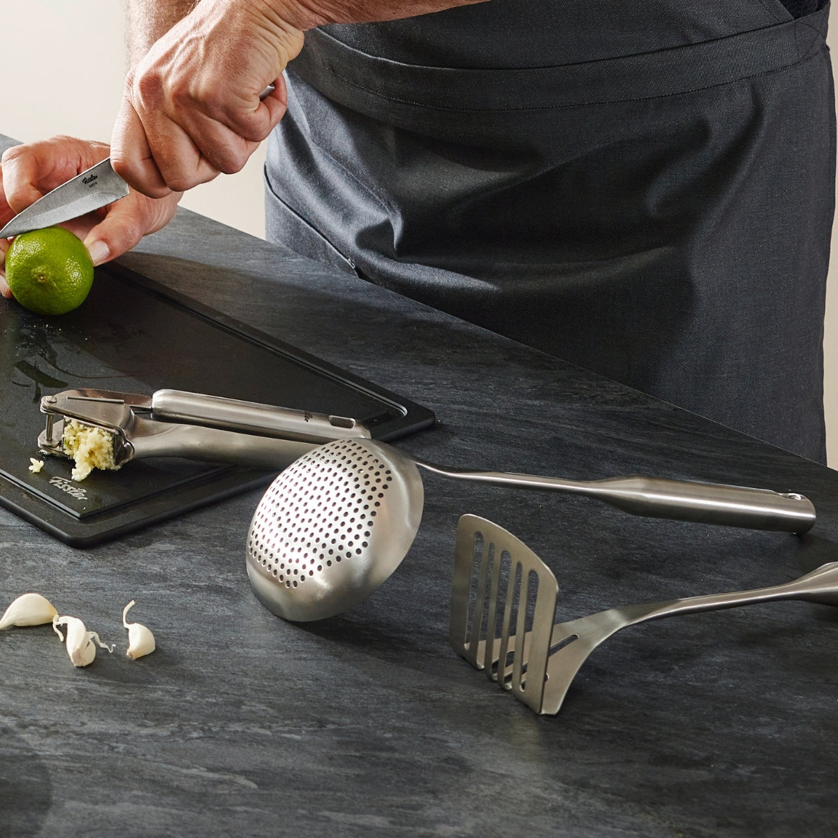 Eine Person in grauer Schürze bereitet auf einer modernen dunklen Fläche neben Knoblauch, einer Presse und dem Fissler Essential® Kartoffelstampfer Lebensmittel zu.