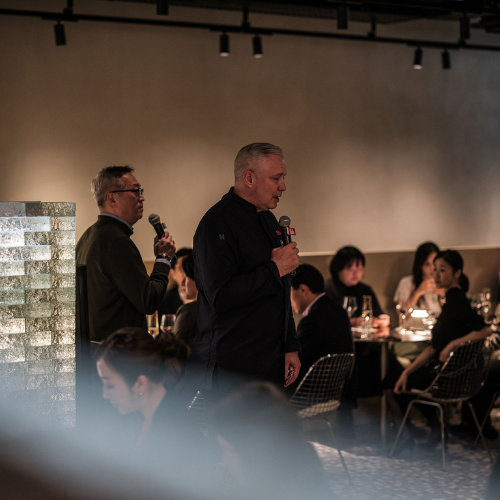 Bei einer Veranstaltung stehen zwei Männer vor Mikrofonen und sprechen zu einem Publikum, das an runden Tischen in einem modernen, sanft beleuchteten Restaurantbereich sitzt. Auf elegant gedeckten Tischen sind die renommierten Edelstahl-Kochgeschirre und Schnellkochtöpfe von Fissler ausgestellt, die das Bekenntnis der Marke zu präziser Technik und zeitlosem Design widerspiegeln.