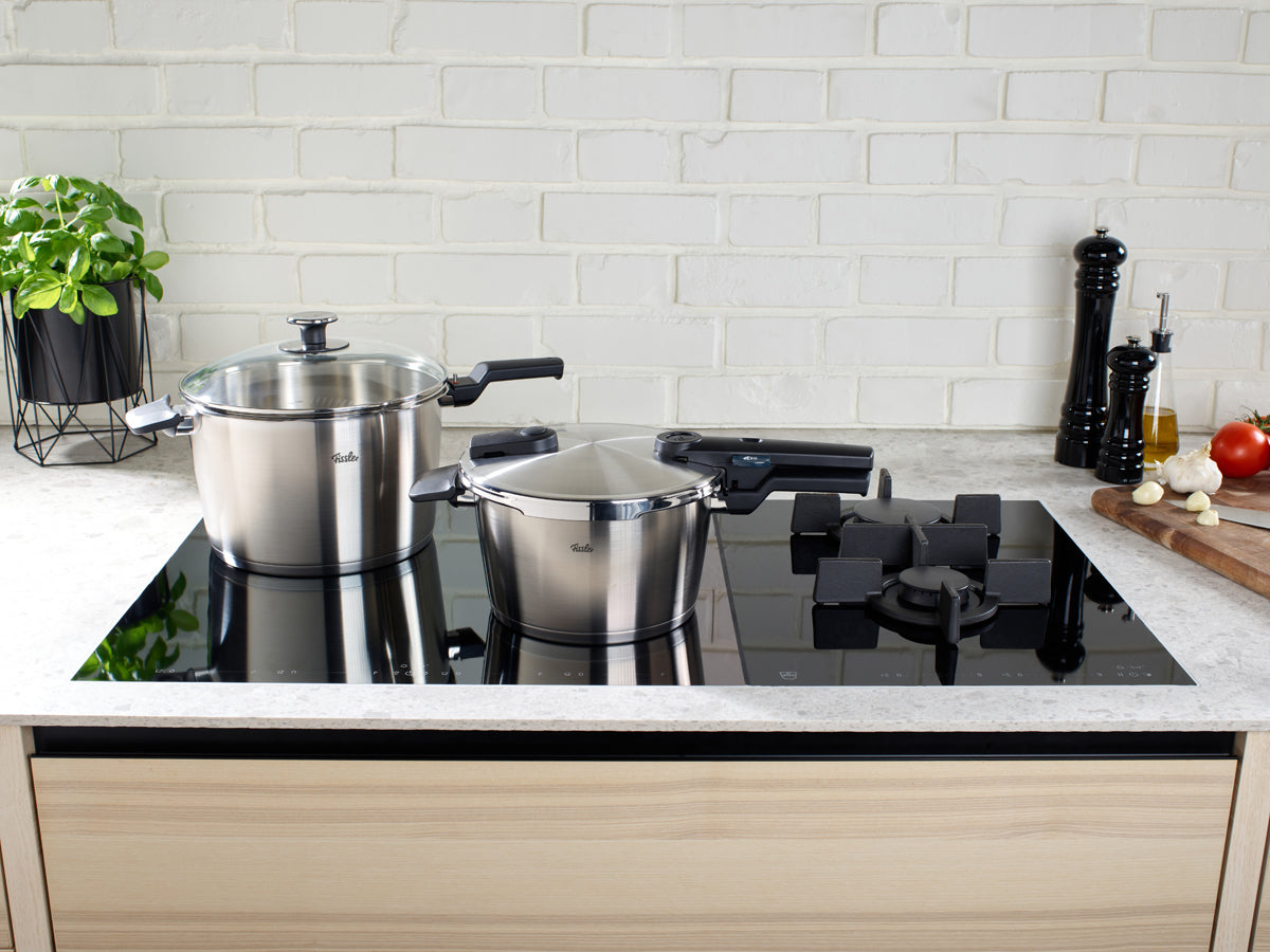 Fissler Vitaquick® Premium 2-tlg. Schnellkochtopf-Set auf dem Kochfeld, mit frischen Kräutern und Küchengeräten in zeitlosem Design.