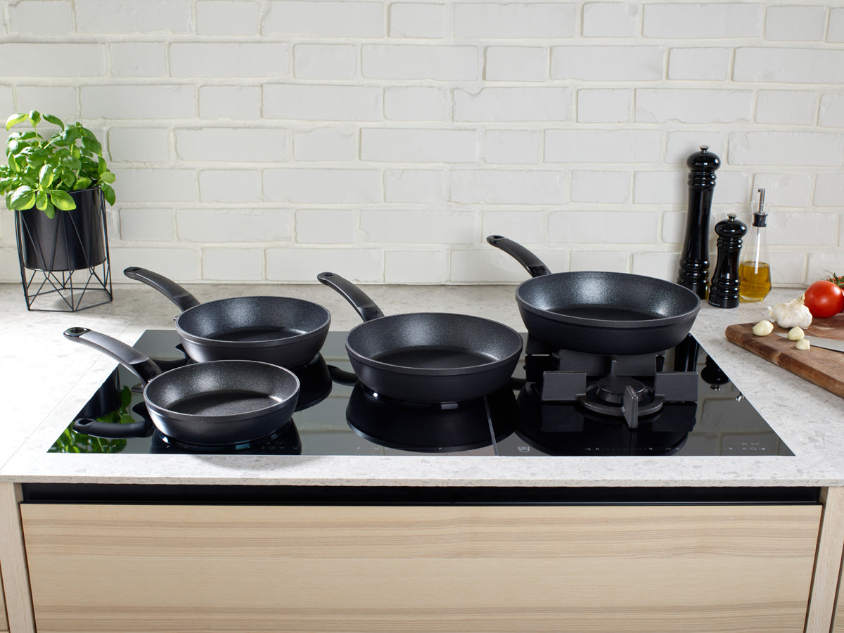 Vier Fissler Adamant® Comfort Antihaft-Pfannen auf einer modernen Herdplatte, mit frischen Kräutern und Gemüse. Präzisionsarbeit in Deutschland.