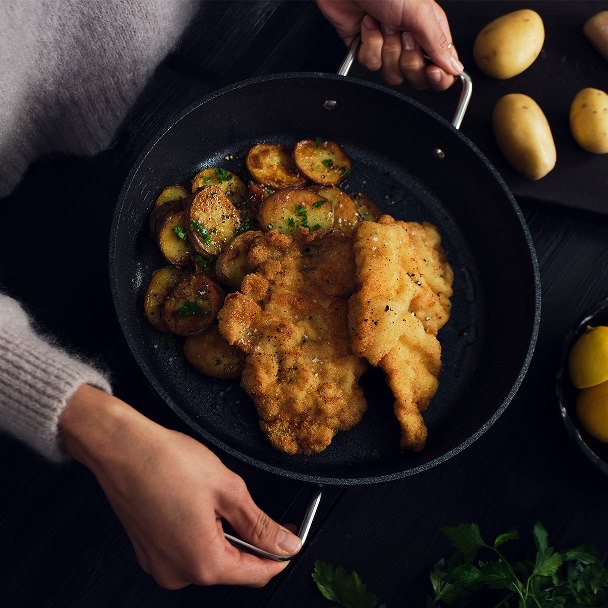 Eine Person präsentiert die Fissler Adamant® Servierpfanne mit zwei perfekt panierten, gebratenen Schnitzeln und goldbraun angebratenen Kartoffelscheiben, die kunstvoll mit Kräutern garniert sind. Die innovative Antihaftbeschichtung und die präzise deutsche Ingenieurskunst – seit 1845 Markenzeichen der Fissler-Tradition – sorgen bei jeder Mahlzeit für müheloses Gelingen. Daneben liegen ganze Kartoffeln und frische Petersilie auf einer eleganten dunklen Tischplatte und unterstreichen die erstklassige