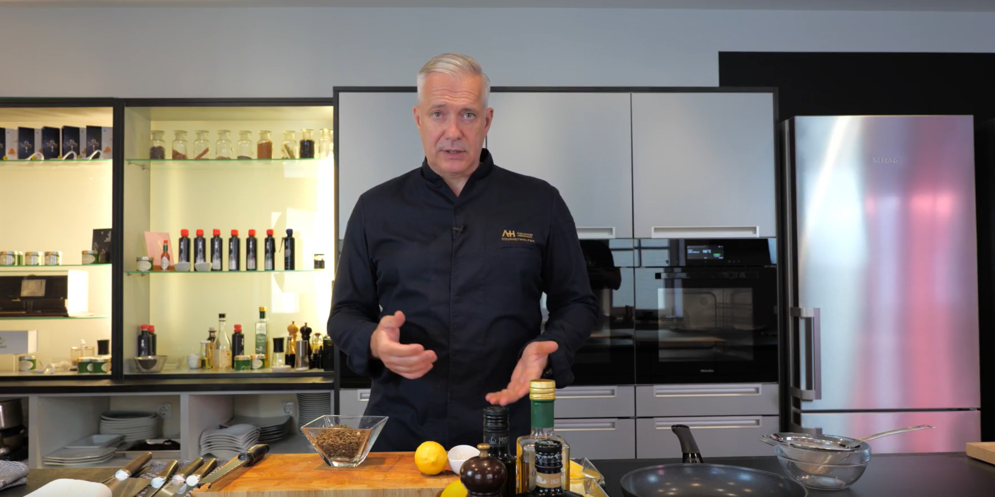 A chef in an elegant kitchen presents Fissler stainless steel cookware alongside fresh ingredients, with shelves and appliances in the background.