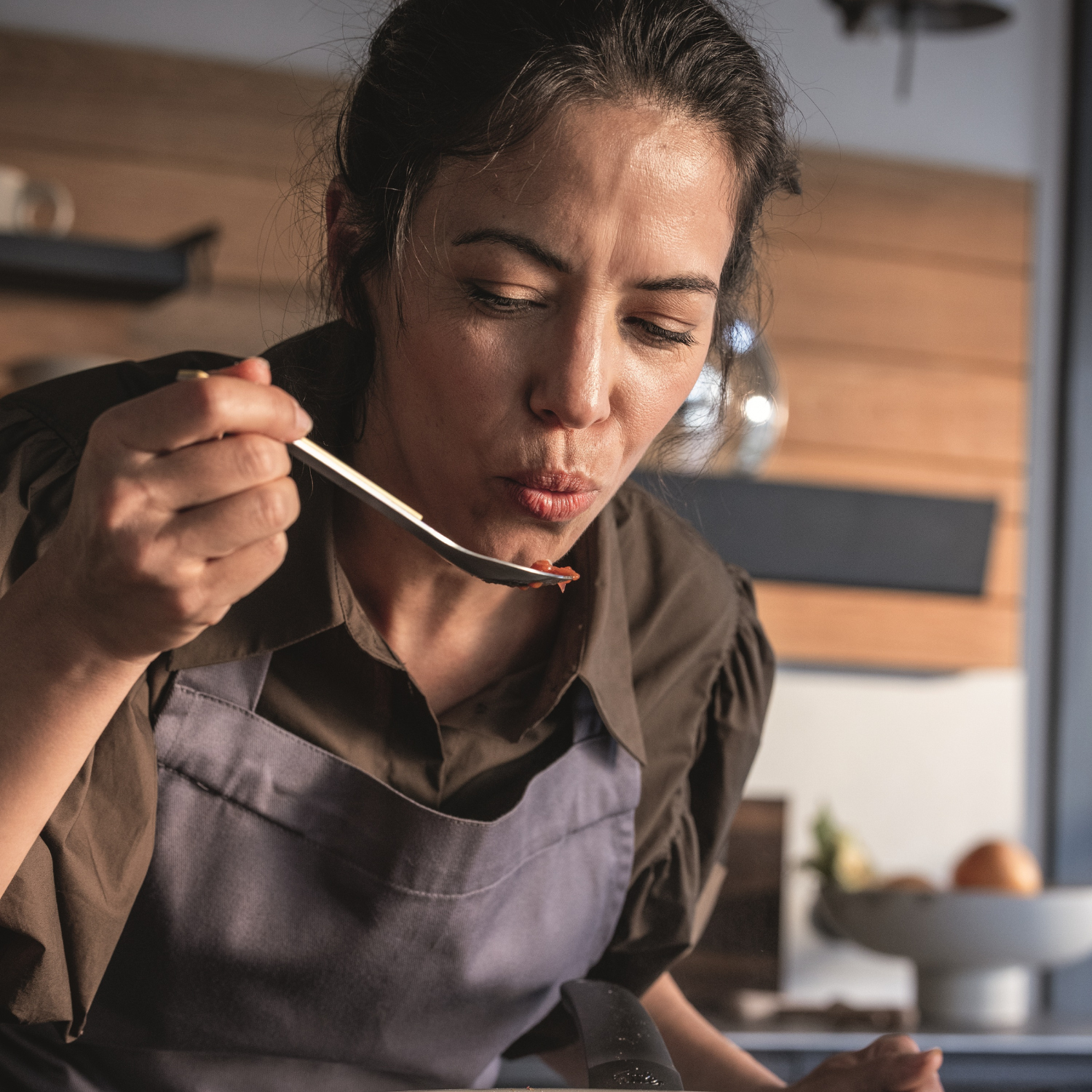 Eine Frau in einer Schürze probiert mit einem Löffel Speisen in einer eleganten, modernen Küche, die mit hochwertigem Fissler-Edelstahl-Kochgeschirr ausgestattet ist. Sie wirkt konzentriert und nachdenklich, während sie den Geschmack bewertet, umgeben von fachmännisch gefertigten Töpfen und Pfannen, die Fisslers berühmte deutsche Handwerkskunst und sein zeitloses Design widerspiegeln.