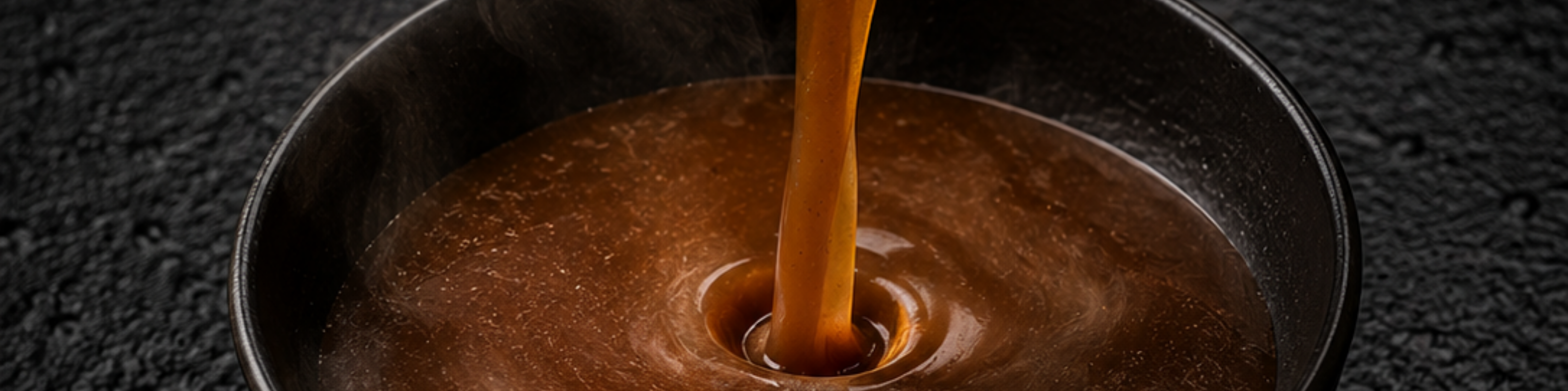 Eine reichhaltige, braune Brühe, die in einem Fissler-Edelstahltopf perfekt gekocht wurde, wird in eine schlichte schwarze Schüssel gegossen, wobei elegante Wellen entstehen und zarte Dampfschwaden aufsteigen – ein Spiegelbild deutscher Präzisionstechnik und zeitlosen Designs.
