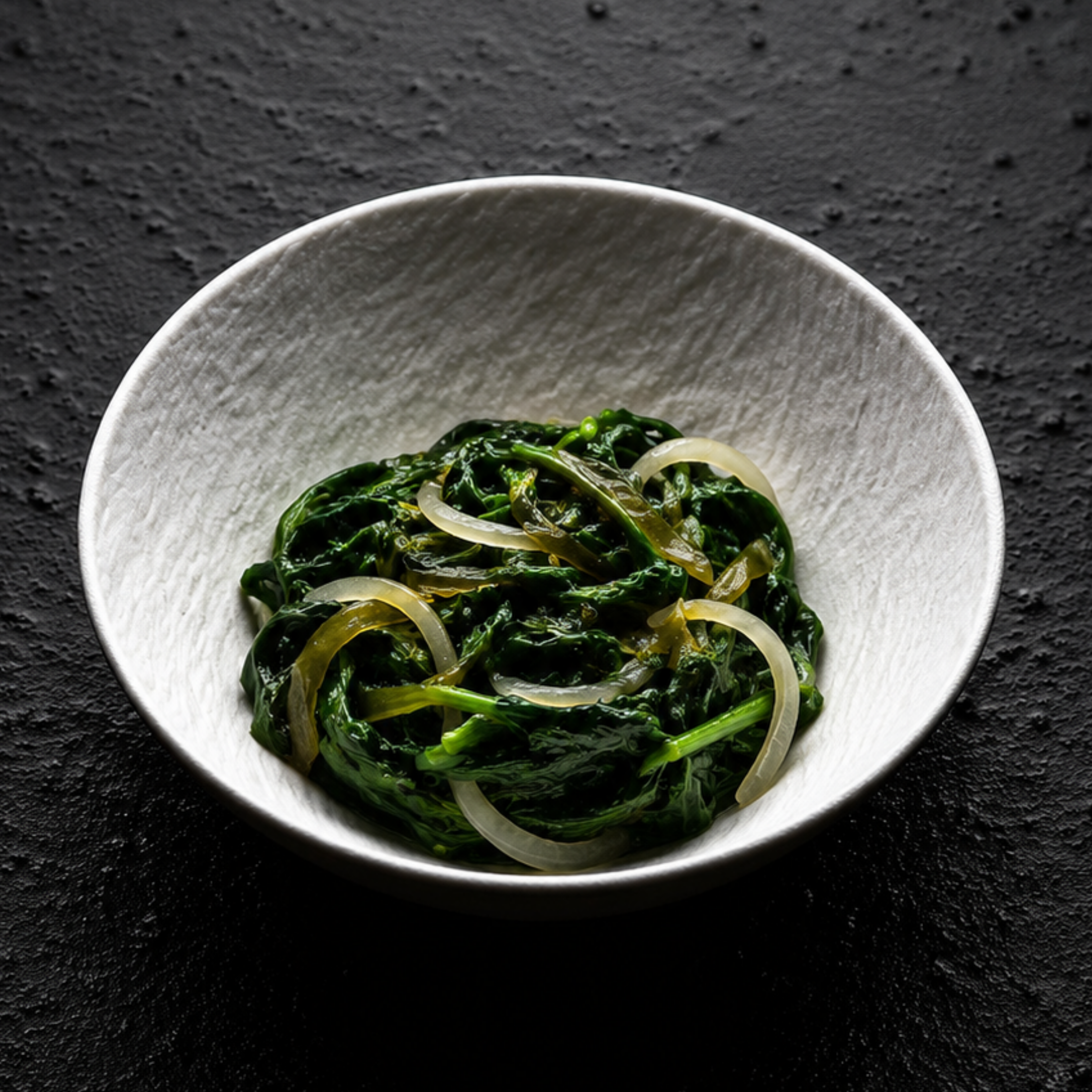 Eine weiße Schüssel präsentiert auf elegante Weise zarten, gegarten Spinat und fein geschnittene Zwiebeln auf einem strukturierten schwarzen Untergrund – eine perfekte Beilage zu Gourmetgerichten, die mit dem präzisionsgefertigten Kochgeschirr von Fissler zubereitet wurden und unsere zeitlose deutsche Handwerkskunst widerspiegeln.