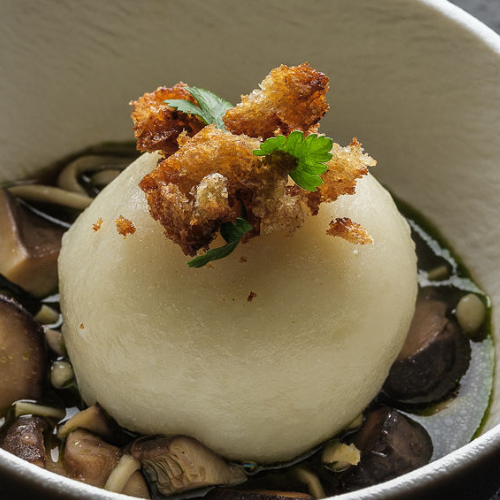 Ein perfekt runder Knödel ruht in einer herzhaften Pilzbrühe, elegant garniert mit goldbraunen, knusprigen Semmelbröseln und einem frischen Petersilienzweig, serviert in einer makellos weißen Schüssel – ein Beispiel für die raffinierte Präsentation, die mit dem präzisionsgefertigten Kochgeschirr von Fissler möglich wird.