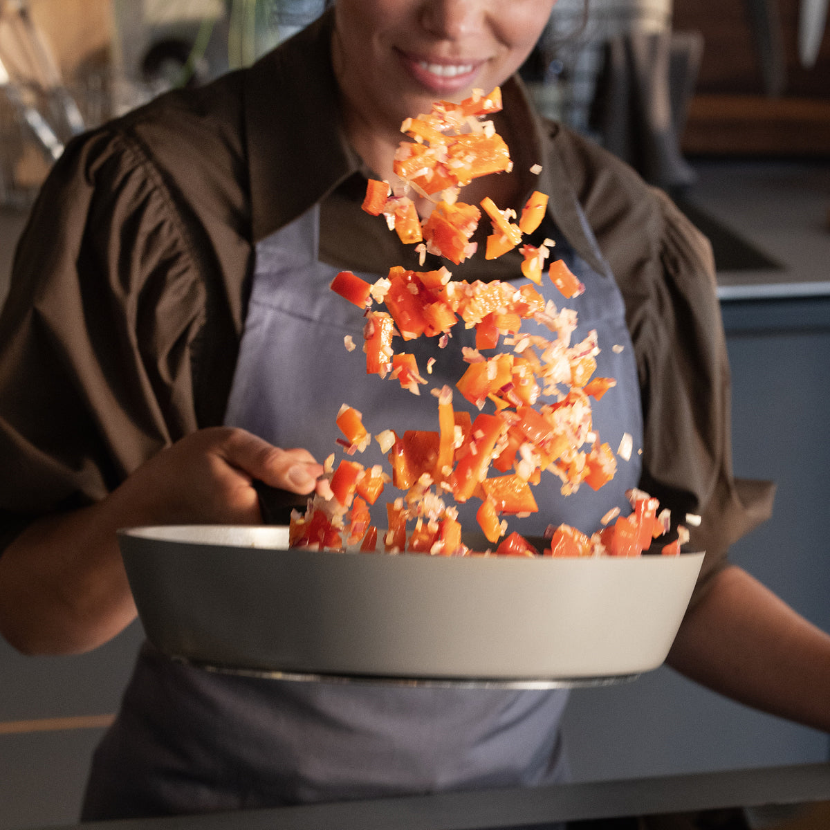 A person in an apron smiles while frying bell peppers and onions in a high-quality Fissler stainless steel pan in a modern kitchen.