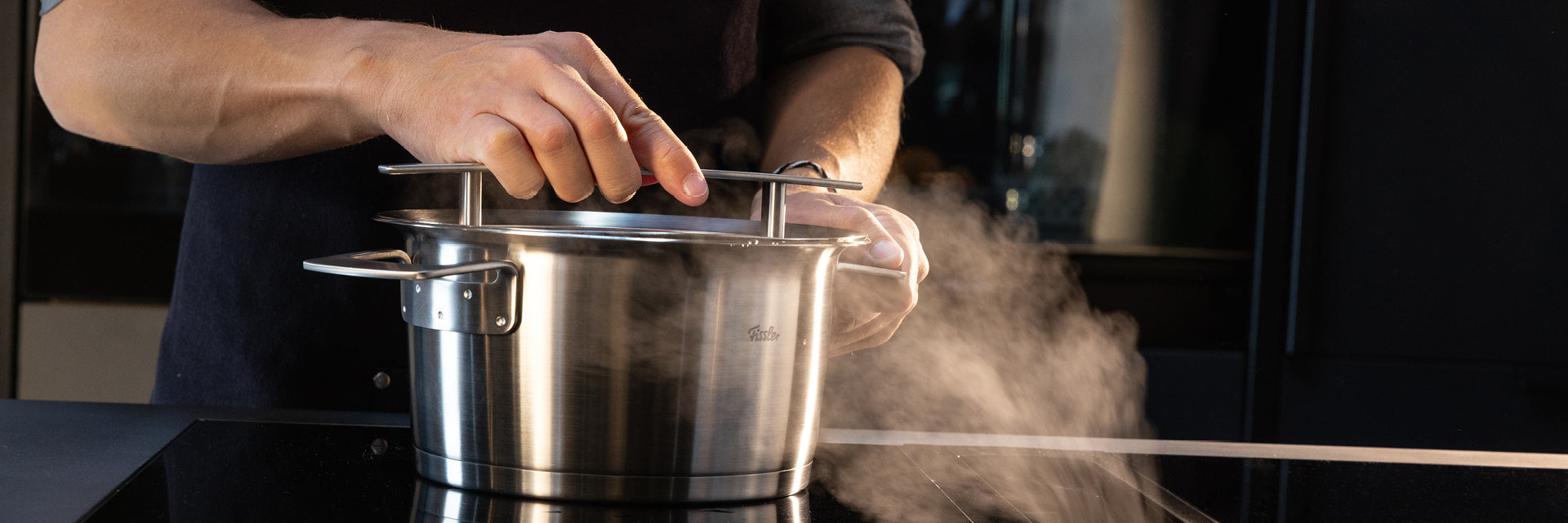 Eine Person hebt den Deckel eines in Deutschland gefertigten Fissler-Edelstahltopfes, während in einer modernen Küche Dampf aufsteigt.
