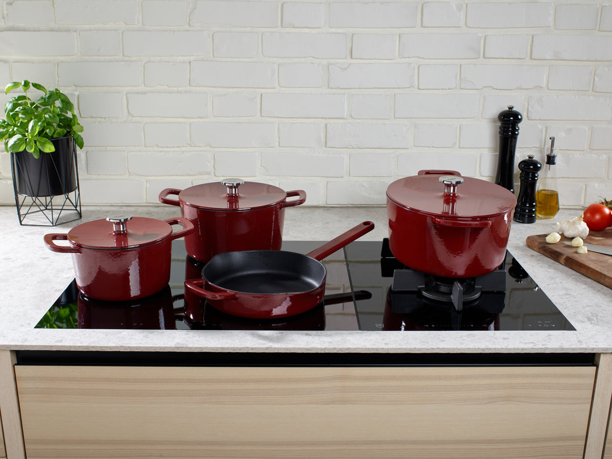 Vier red Fissler Moments cast-iron roasters with Lid on an induction hob, surrounded by fresh ingredients - Made in Germany.