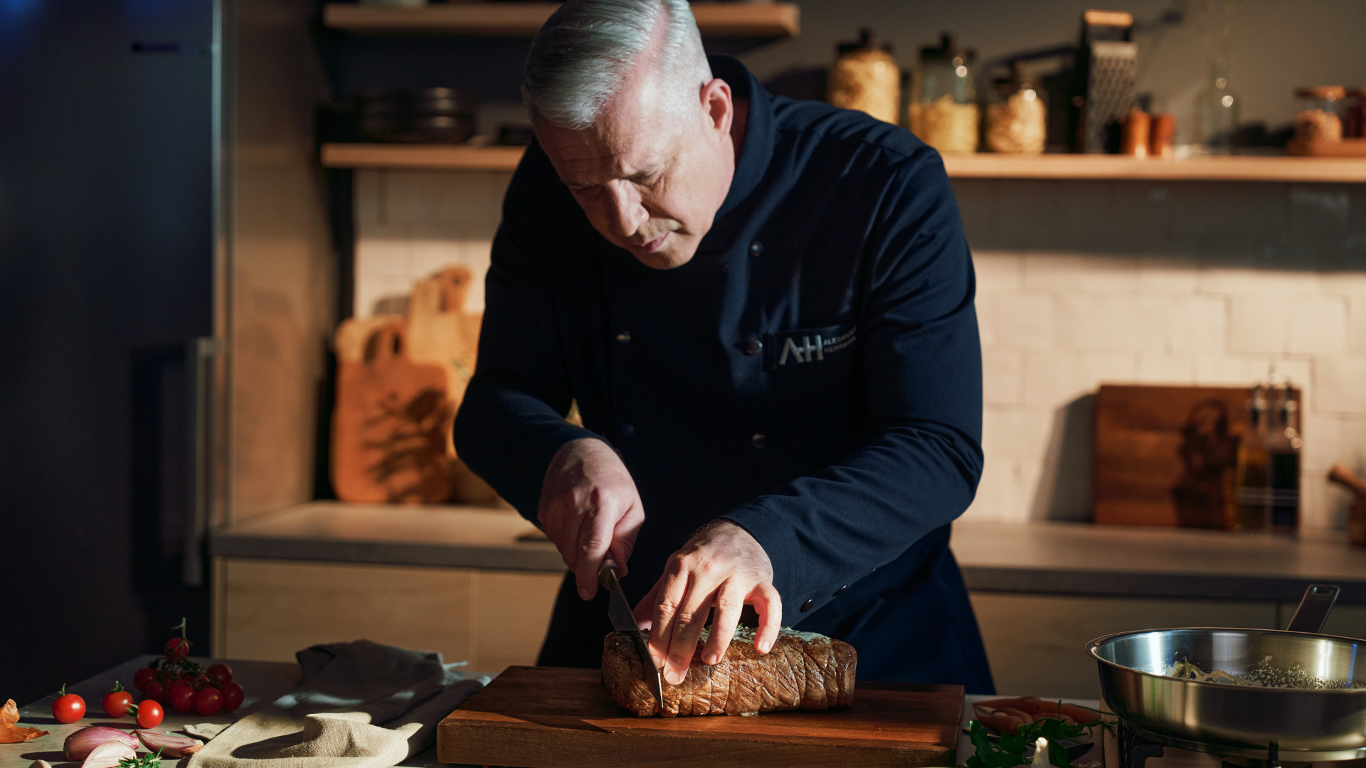 Ein Koch mit grauem Haar in dunkler Uniform schneidet in einer eleganten, modernen Küche mit Fissler-Kochgeschirr Braten auf einem Holzbrett.