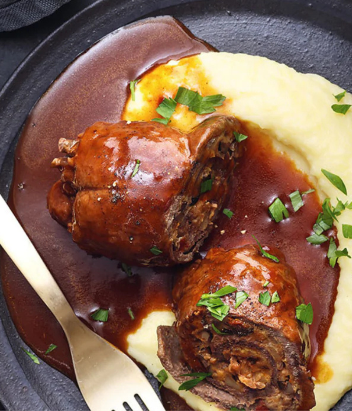 Zarte geschmorte Fleischroulade in brauner Sauce auf Kartoffelpüree, Kräutergarnitur; serviert auf einem dunklen Teller - mit Fissler-Präzision zubereitet.