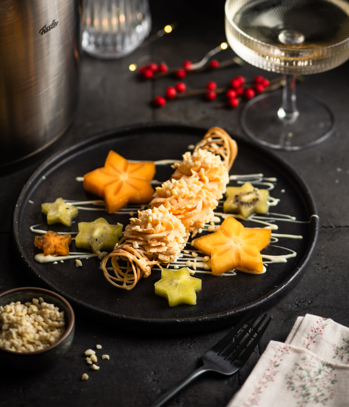 Ein mit Präzision zubereitetes Dessert mit Gebäckröllchen, Fruchtsternen und Sauce - hergestellt mit Fissler-Kochgeschirr. Champagner und Dekor dahinter.