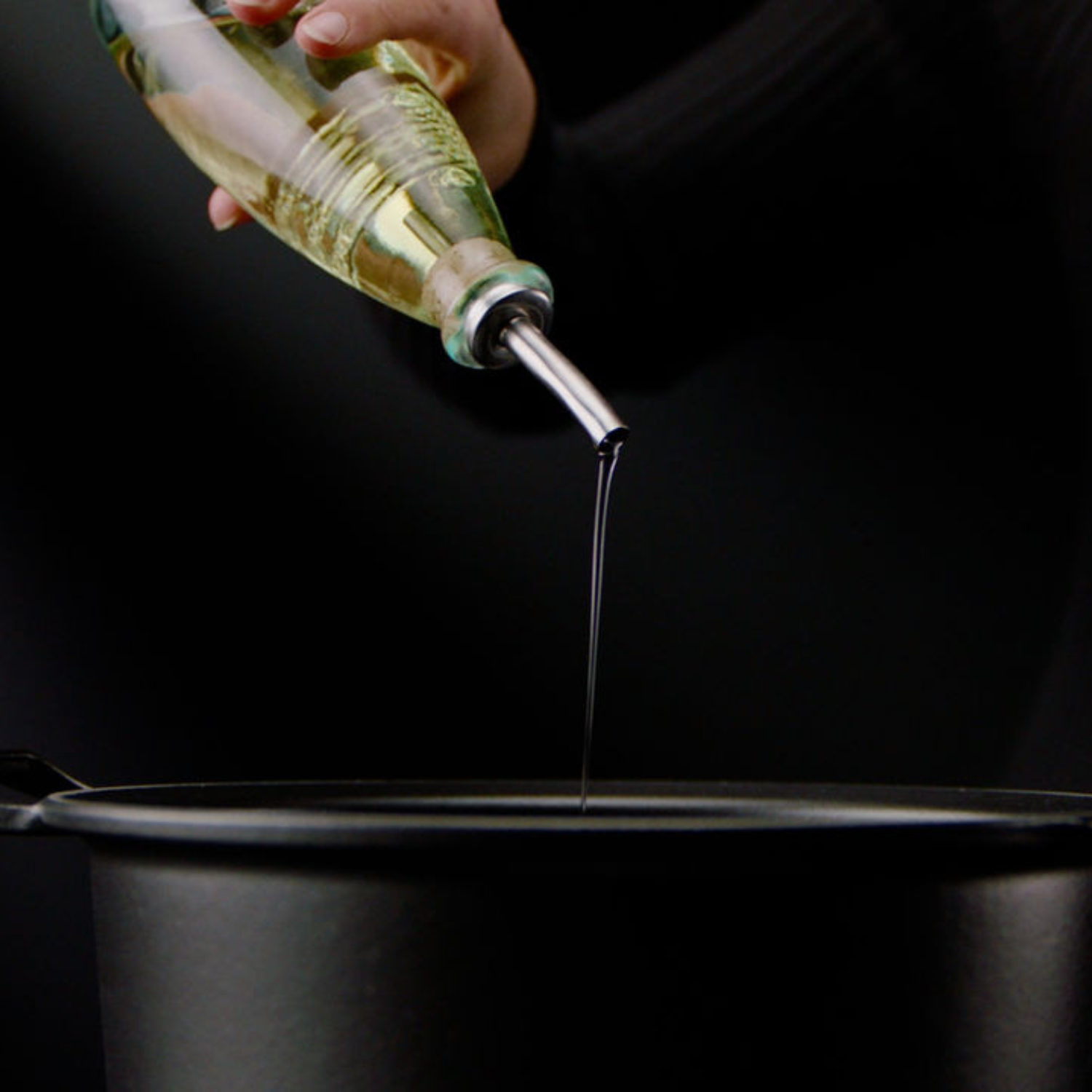 Eine Hand gießt Öl aus einer Glasflasche in einen hochwertigen schwarzen Fissler-Topf, der zeitloses deutsches Design und präzise Handwerkskunst zeigt.