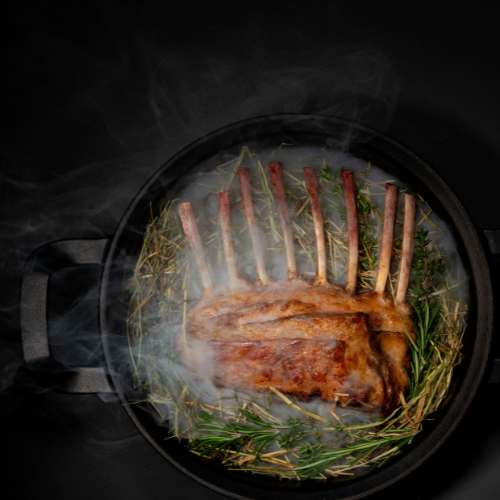 Saftiges Lammfleisch auf frischen Kräutern in einer schwarzen Fissler-Bratpfanne, präzise gegart in wirbelndem Dampf, vor dunklem Hintergrund.