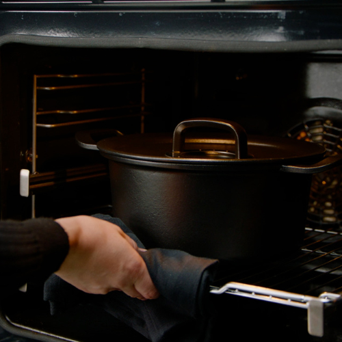 Eine Person stellt einen gusseisernen Dutch Oven mit Deckel von Fissler auf einen Ofenrost und benutzt zur Sicherheit einen Topflappen - zeitloses deutsches Design.