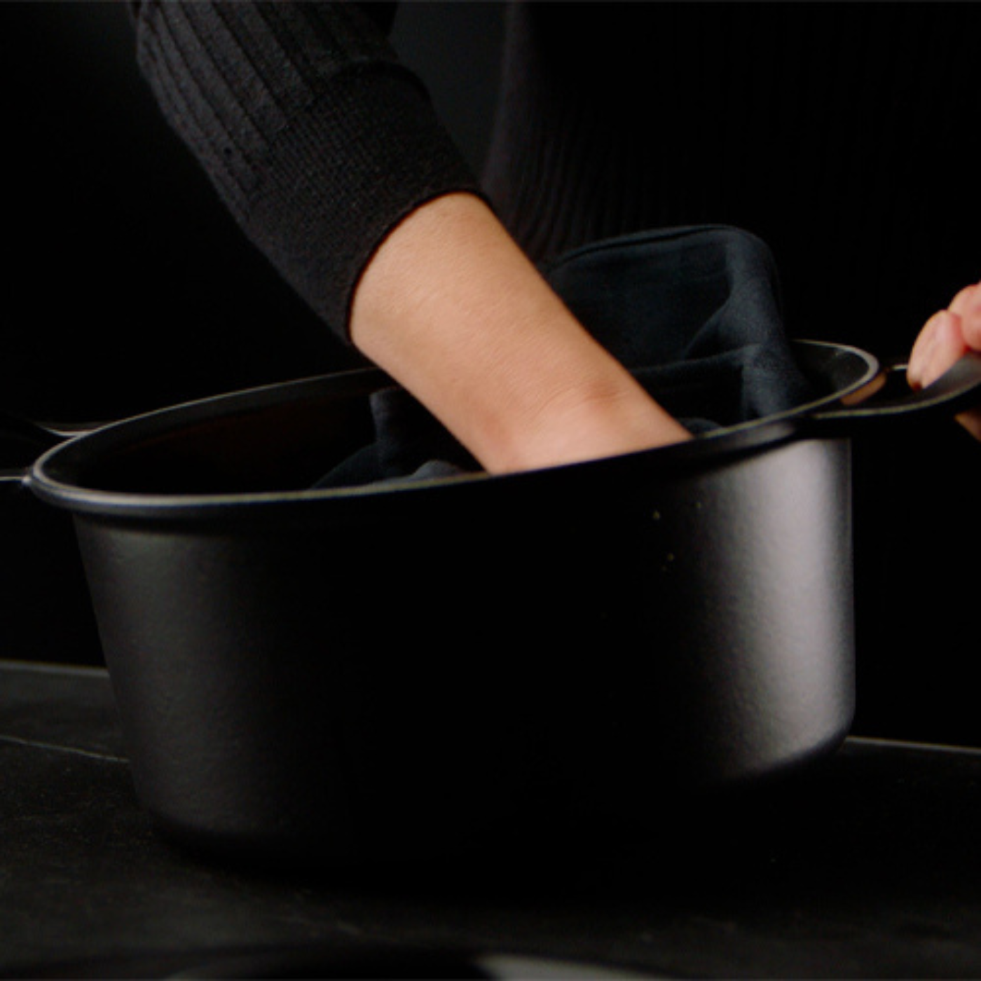 Eine Person legt ein Tuch in einen hochwertigen schwarzen Fissler-Topf, der mit Präzision auf einer glatten dunklen Oberfläche und einem dunklen Hintergrund gefertigt wurde.