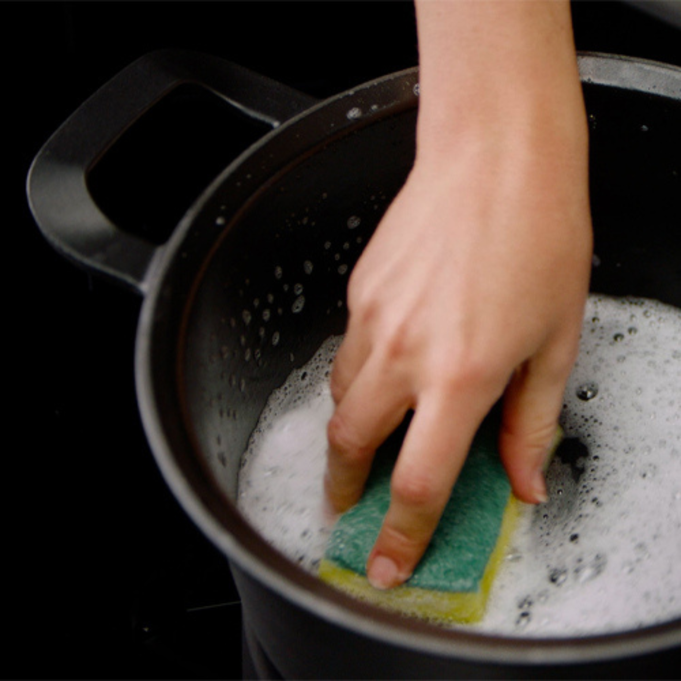 Eine Hand reinigt einen hochwertigen schwarzen Fissler-Topf mit einem grün-gelben Schwamm und unterstreicht damit die Langlebigkeit und Präzision deutscher Produktion.