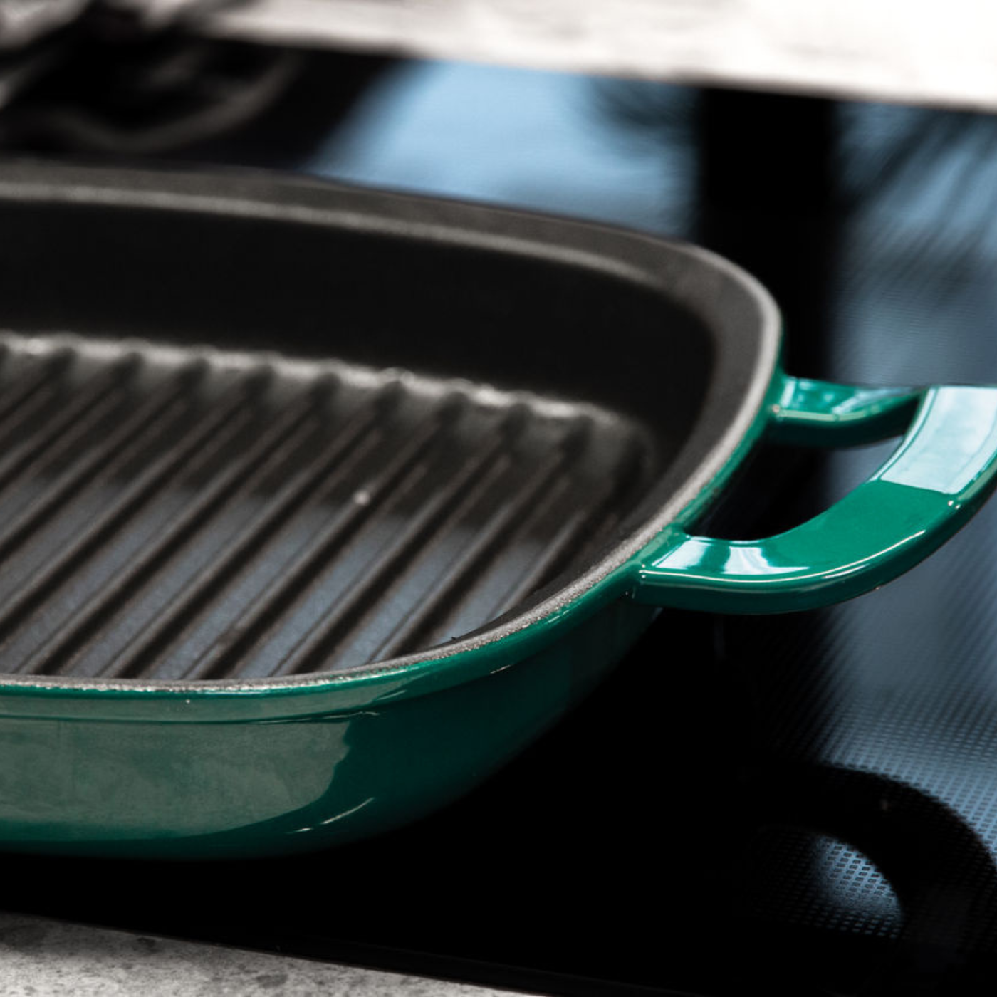 Eine Nahaufnahme einer grünen gusseisernen Grillpfanne von Fissler mit präzisen Rillen, die auf einer glatten schwarzen Glasplatte präsentiert wird.