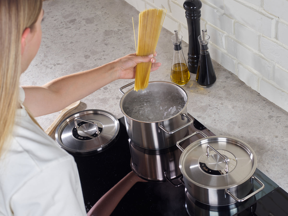 A person cooks spaghetti over boiling water with two Fissler Pure Collection pots with Metal Lid on the counter, made with precision craftsmanship.