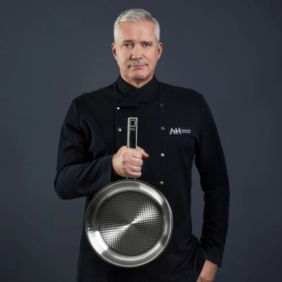 Ein vornehmer Koch in Schwarz hält eine Fissler-Edelstahlbratpfanne und präsentiert deutsche Präzision vor einem dunklen Hintergrund.