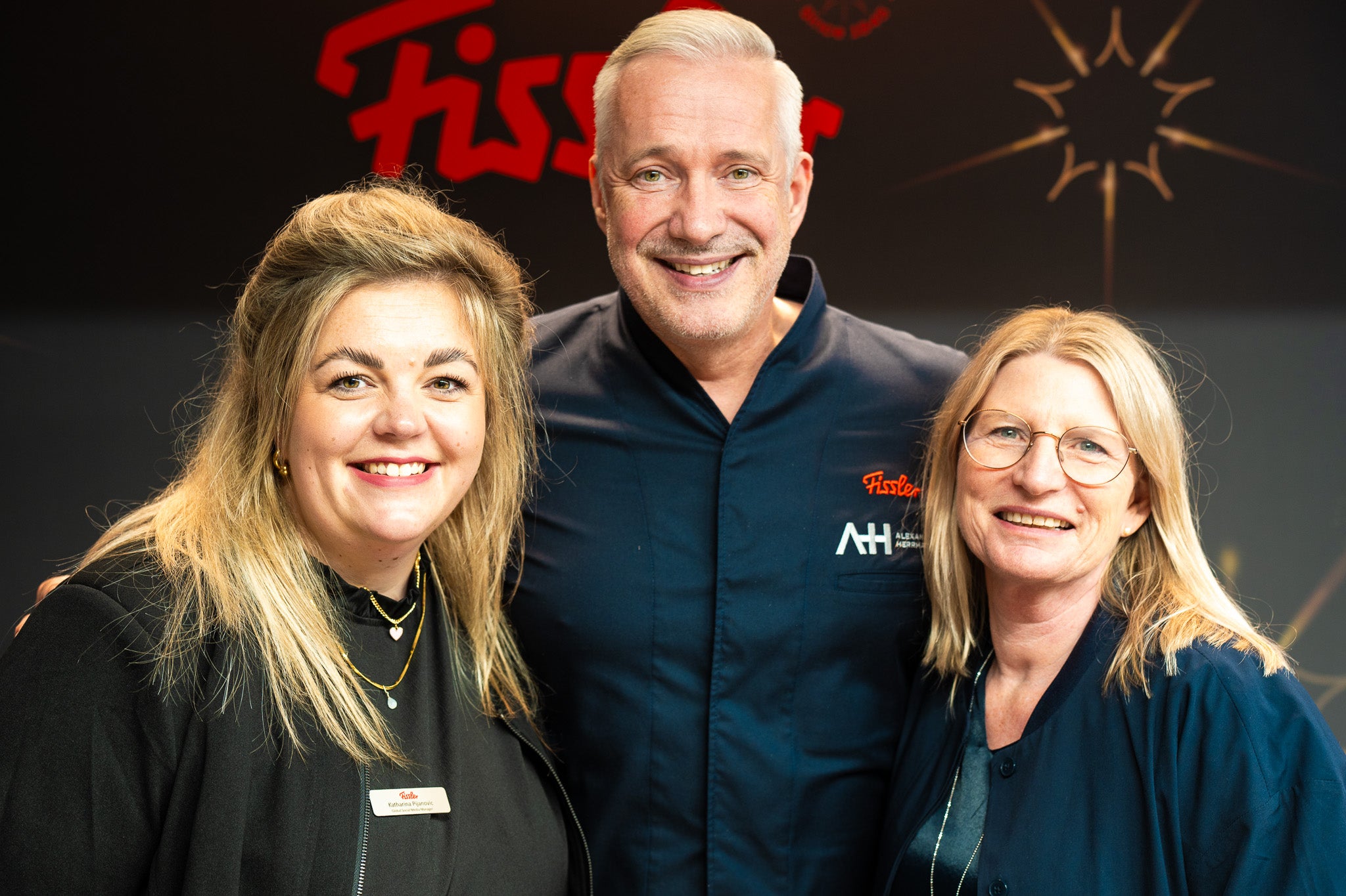 Drei Erwachsene, zwei Frauen und ein Chefkoch, lächeln in der Küche. Das Fissler-Logo und der Sternenhimmel unterstreichen die Tradition unseres in Deutschland hergestellten Premium-Kochgeschirrs.