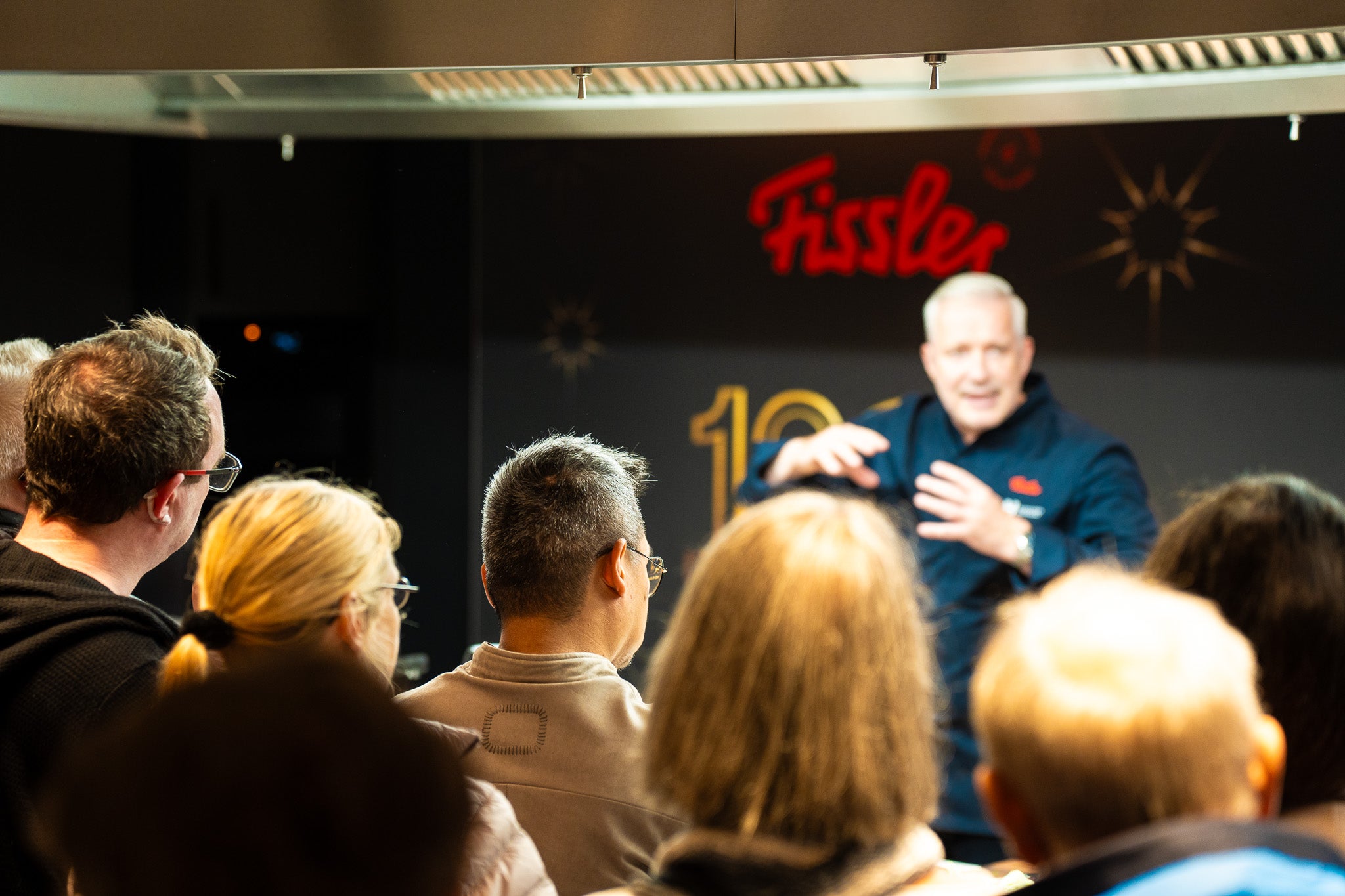 Ein Chefkoch präsentiert sich dem Publikum in einer modernen Küche, mit Fissler-Schild und Kochgeschirr, die für deutsche Präzision seit 1845 stehen.