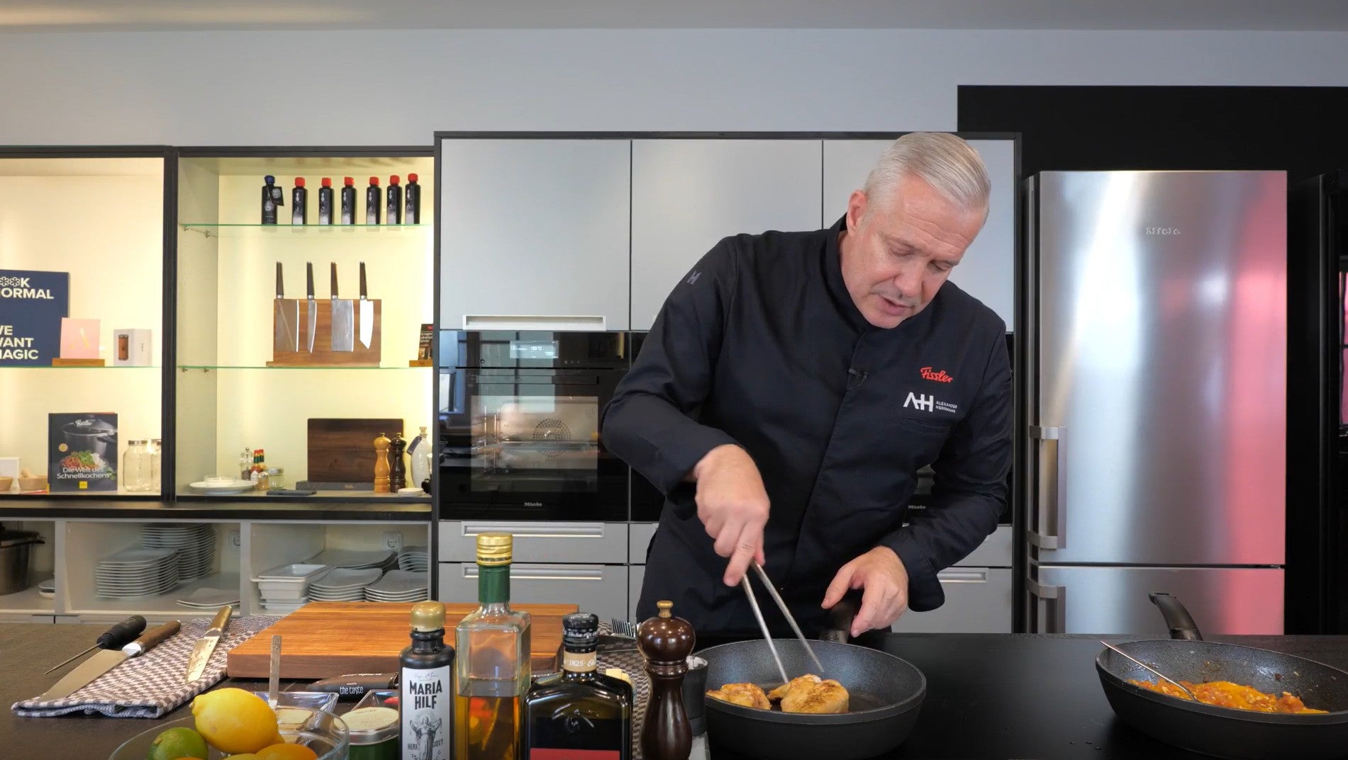 A chef cooks in a modern kitchen with Fissler stainless steel cookware. Ingredients and spices are laid out on the counter next to it.