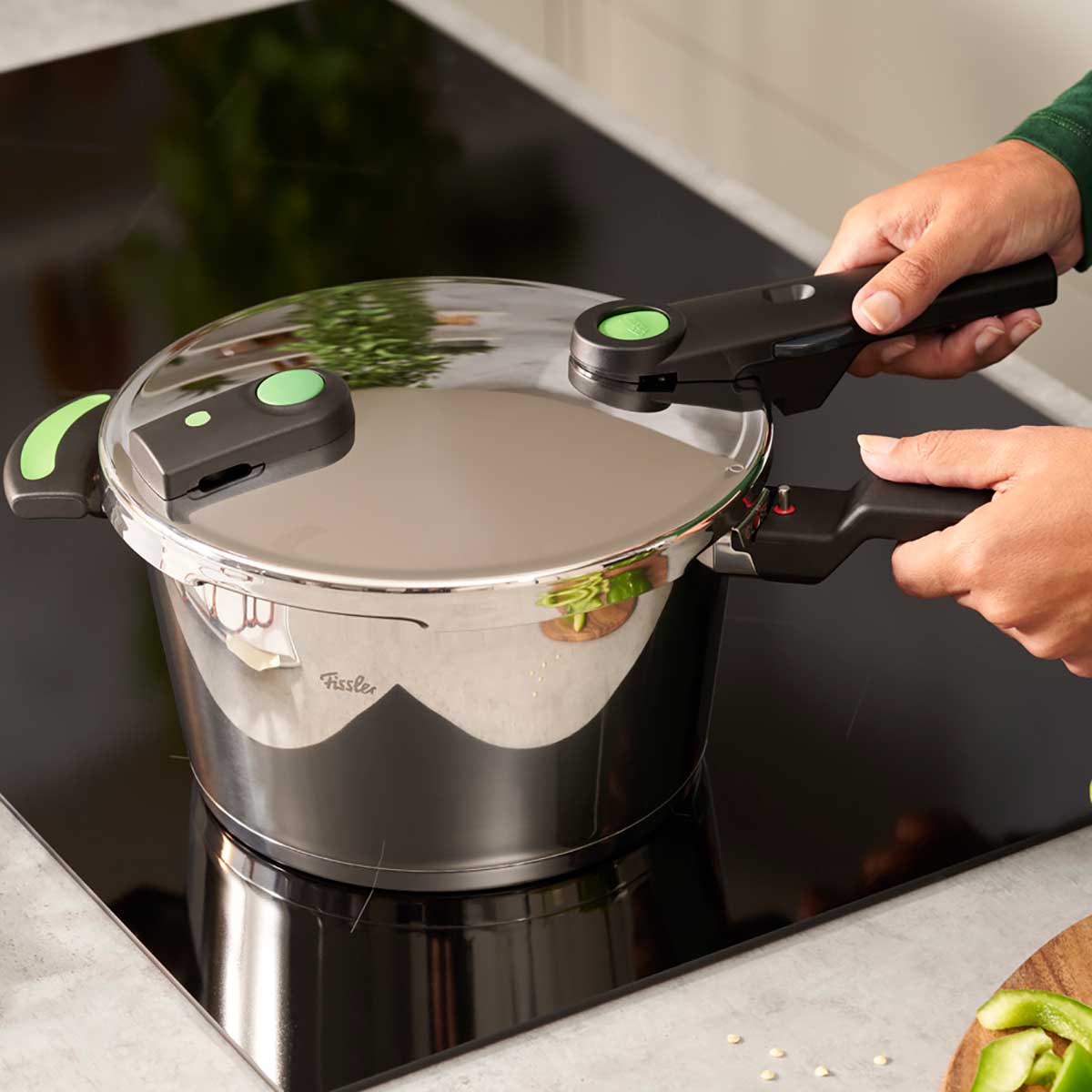 A person attaches the lid of a Fissler Vitaquick® Green pressure cooker with black and green handles to a stovetop in a sleek, modern kitchen.