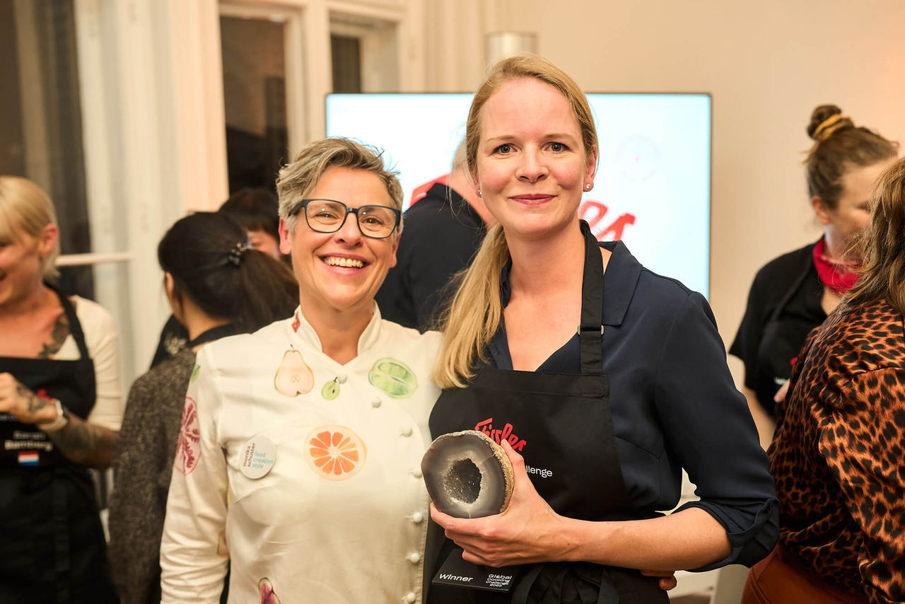 Zwei Frauen lächeln bei einer Veranstaltung; die eine trägt eine Fissler-Schürze und hält einen hochwertigen deutschen Kochgeschirrdeckel, während sich im Hintergrund die Gäste unterhalten.