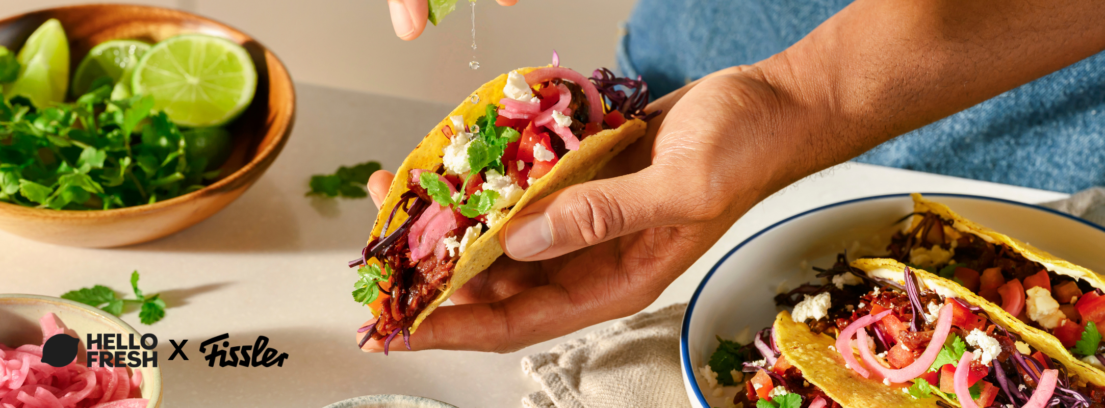 Eine Hand presst frische Limette über farbenfrohe Tacos, die mit Gemüse und Käse gefüllt sind; daneben liegen zusätzliche Limettenschnitze und Koriander. Die Szene spielt sich auf hochwertigem Fissler-Edelstahlgeschirr ab, das deutsche Präzisionstechnik und zeitloses Design widerspiegelt. In der unteren linken Ecke sind „Hello Fresh x Fissler“-Logos zu sehen, die die hohe Qualität der Küchenausstattung unterstreichen.