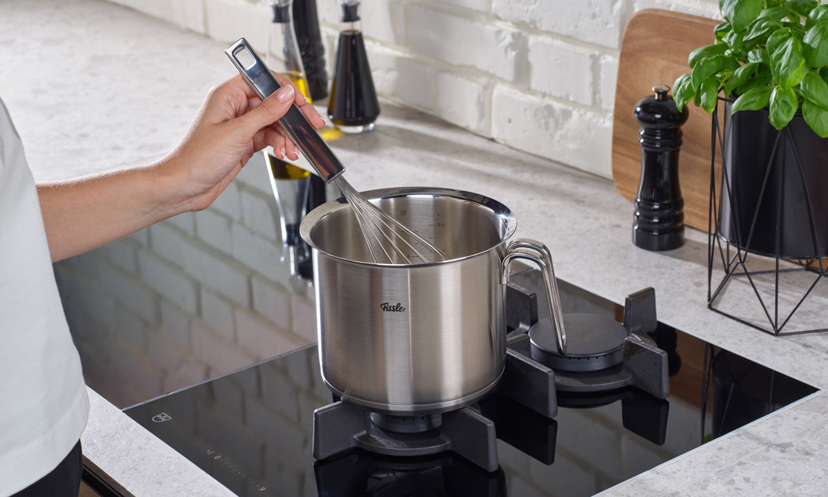 A person uses a metal whisk with the Fissler Original-Profi Collection® milk pot 16cm on a modern gas stove in a bright kitchen.