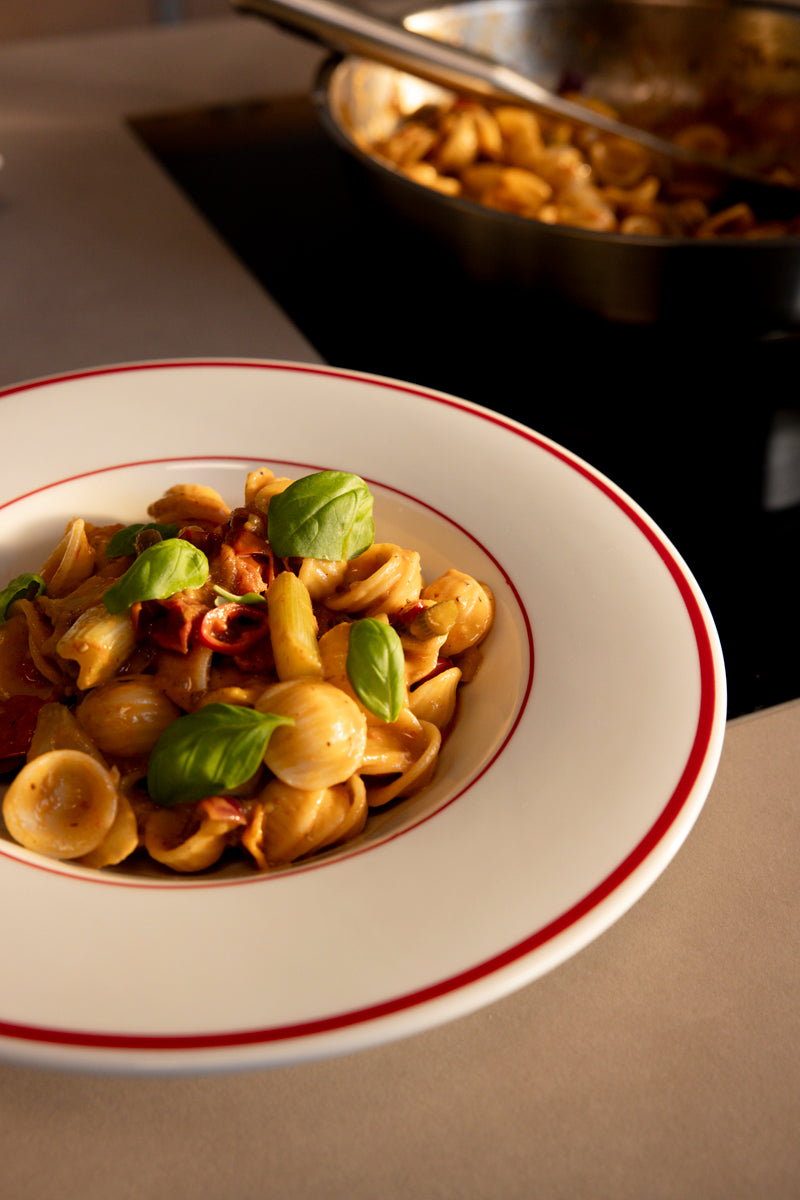 Orecchiette-Nudeln in Tomatensauce mit Basilikum auf einem Teller; im Hintergrund weitere Nudeln in einer Fissler-Pfanne auf dem Herd.