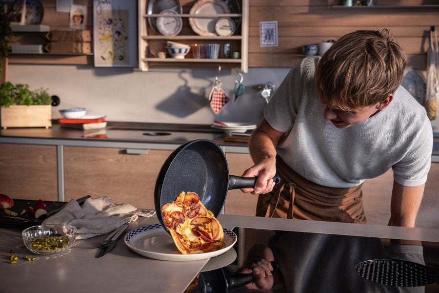 Ein Koch im hellen Hemd benutzt eine Fissler-Pfanne, um in einer modernen Küche mit Holzschränken ein Omelett auf einem Teller zu servieren.