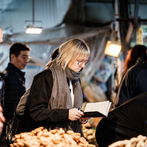 Eine Person mit blonden Haaren und Brille schreibt in ein Notizbuch, während sie an einem Marktstand mit einer Auswahl an frischen Pilzen steht – ein Bild, das die Inspiration für außergewöhnliche Kochkunst vermittelt. Andere Marktbesucher und warmes Licht unterstreichen die lebendige Atmosphäre und spiegeln die Liebe zu hochwertigen Zutaten wider, die perfekt zu Fisslers präzise gefertigtem Edelstahl-Kochgeschirr und den zeitlosen Küchenaccessoires passt.