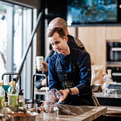 Ein Koch in blauer Uniform und Schürze reinigt Fissler-Edelstahltöpfe und -Pfannen an einer modernen Küchentheke, umgeben von präzise gefertigten Küchenutensilien und hochwertigen Zutaten. Im Hintergrund steht eine weitere Person mit einem Getränk in der Hand, was die einladende Atmosphäre unterstreicht, die Fisslers zeitloses Kochgeschirr in jede heimische Küche bringt.
