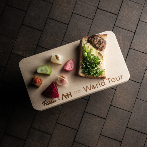 Auf einem Holzbrett mit der Aufschrift „World Tour“ ist eine sorgfältig angerichtete Scheibe Brot mit frischem Blattgemüse, verschiedenen eingelegten Gemüsesorten und einem kleinen Stück Sushi zu sehen, alles auf einer eleganten, dunklen Fliesenoberfläche – eine einladende Präsentation kulinarischer Kreationen, die mit den präzisionsgefertigten Küchenutensilien von Fissler zubereitet wurden.