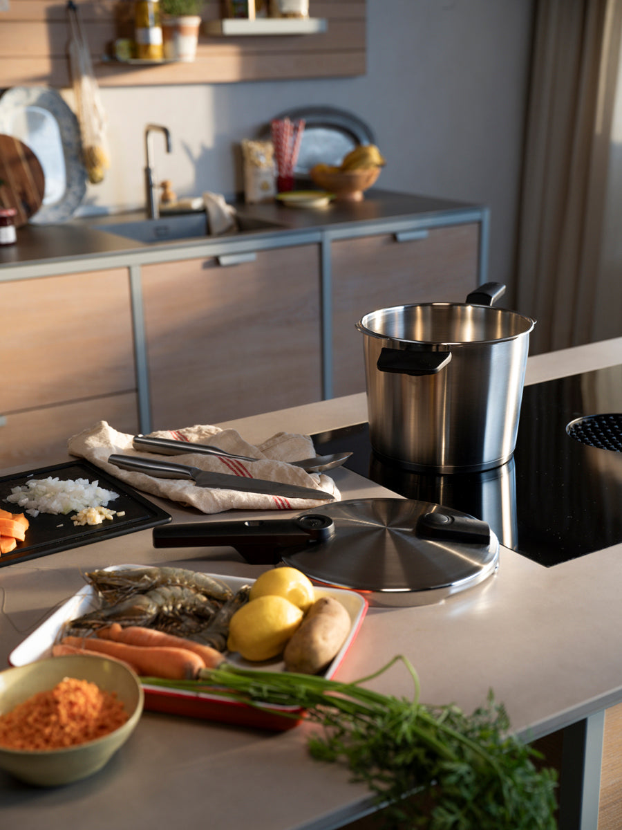 A sophisticated kitchen counter showcases fresh vegetables, prawns, and a bowl of grains artfully arranged beside the premium Fissler Vitaquick® pressure cooker with steamer insert and included cookbook. Chopped onions, a fine kitchen knife, and soft cotton towels enhance the scene, all set within an elegant, modern kitchen—reflecting Fissler’s heritage of precision engineering and innovative cookware “Made in Germany.” Perfect for preparing fast, delicious meals with our high-quality stainless steel pots