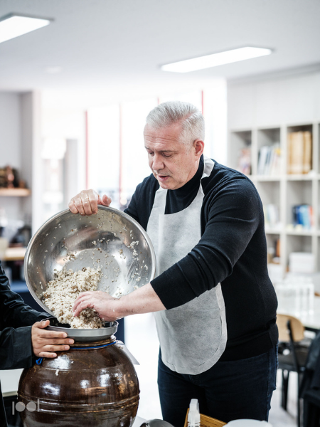 In einer hellen, modernen Küche schüttet ein Mann in einer strahlend weißen Schürze eine Schüssel Reis oder eine Getreidemischung in ein großes Keramikgefäß. Auf der Arbeitsplatte sind elegantes Fissler-Edelstahlgeschirr und hochwertiges, in Deutschland hergestelltes Küchenzubehör zu sehen, das höchste Präzision und zeitloses Design widerspiegelt.