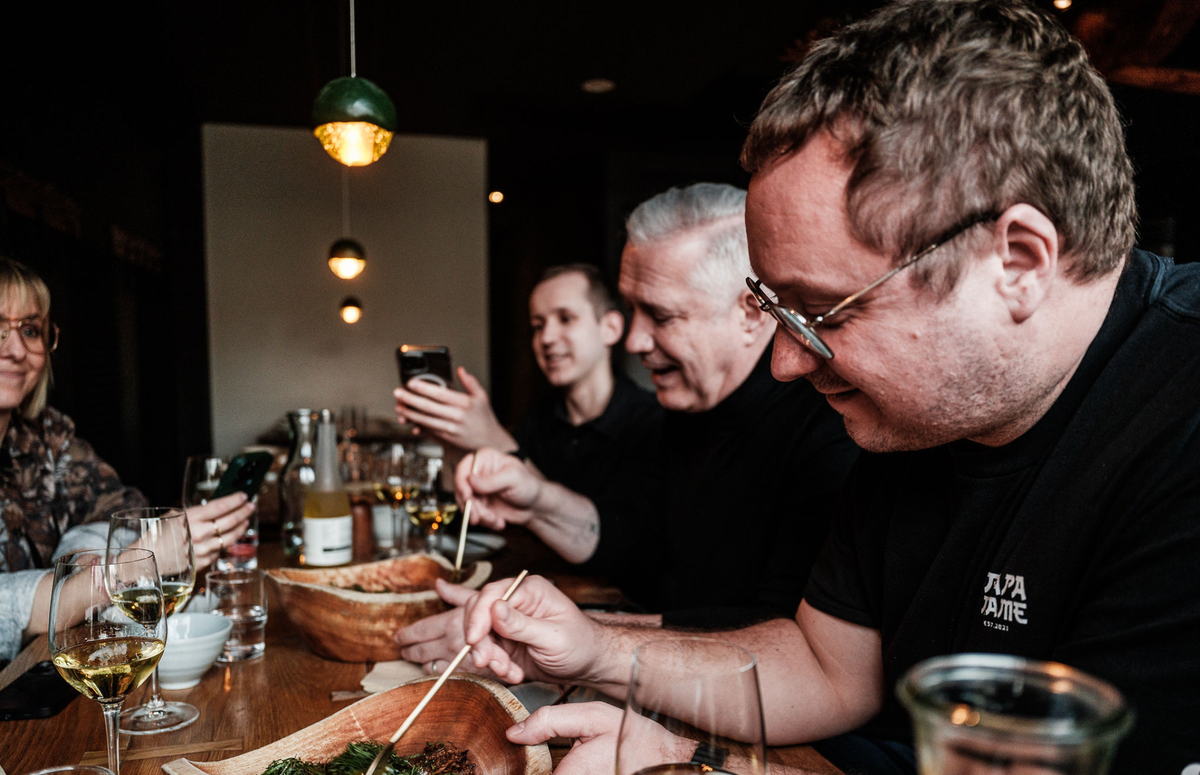 Four people gather around a restaurant table, enjoying expertly prepared dishes with chopsticks. Two guests capture the moment on their phones, while Fissler’s elegant stainless steel cookware and serving accessories are featured among the wine glasses and plates on the wooden table—a reflection of precision engineering and timeless German design enhancing every dining experience.