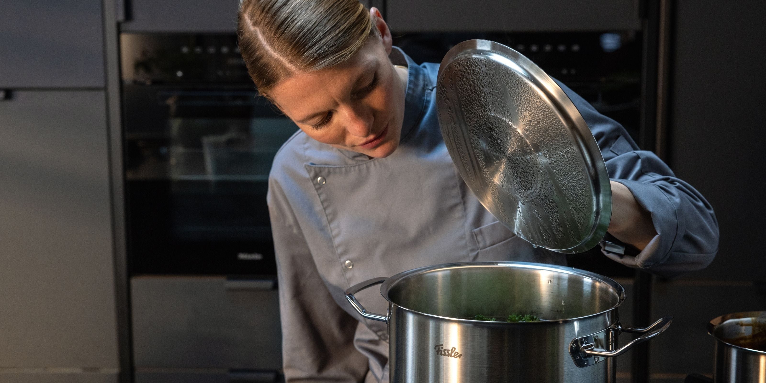 Ein Koch in grauer Uniform hebt den Deckel eines Fissler-Edelstahltopfes und zeigt damit deutsche Präzision und zeitloses Design in der Küche.