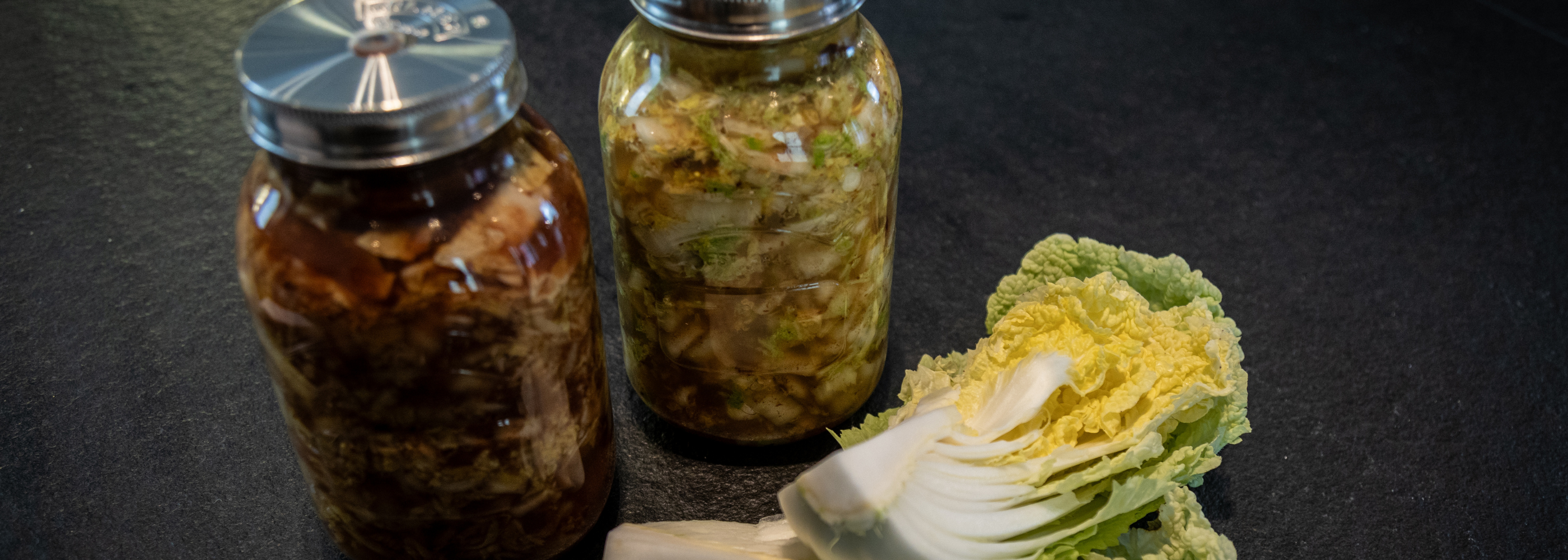 Zwei mit hausgemachtem Kimchi gefüllte Gläser stehen auf einer dunklen Arbeitsplatte neben einem halbierten Kopf frischem Chinakohl und vermitteln einen Eindruck von Präzision und Frische. Diese Inszenierung unterstreicht die Bedeutung hochwertiger Küchenutensilien – wie beispielsweise des Edelstahl-Kochgeschirrs oder des Zubehörs von Fissler – bei der Zubereitung und Aufbewahrung köstlicher hausgemachter Gerichte.
