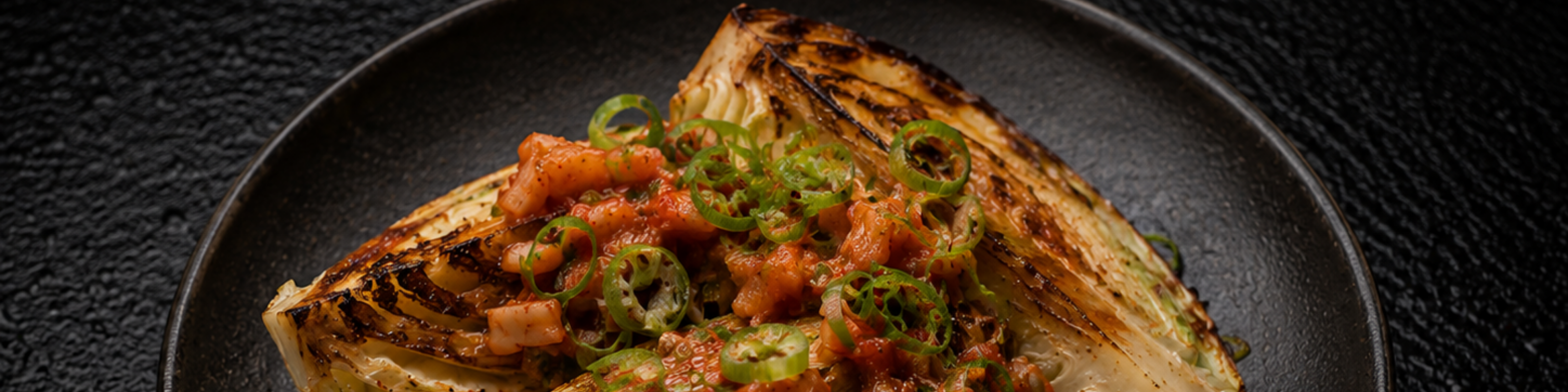 Ein gegrilltes Kohlstück, garniert mit Tomatenwürfeln und in Scheiben geschnittenen grünen Chilischoten, elegant auf einem dunklen Teller vor einem strukturierten schwarzen Hintergrund angerichtet. Das Gericht, das mit hochwertigem Fissler-Kochgeschirr zubereitet wurde, spiegelt das Bekenntnis der Marke zu präziser Technik und zeitlosem deutschem Design wider.
