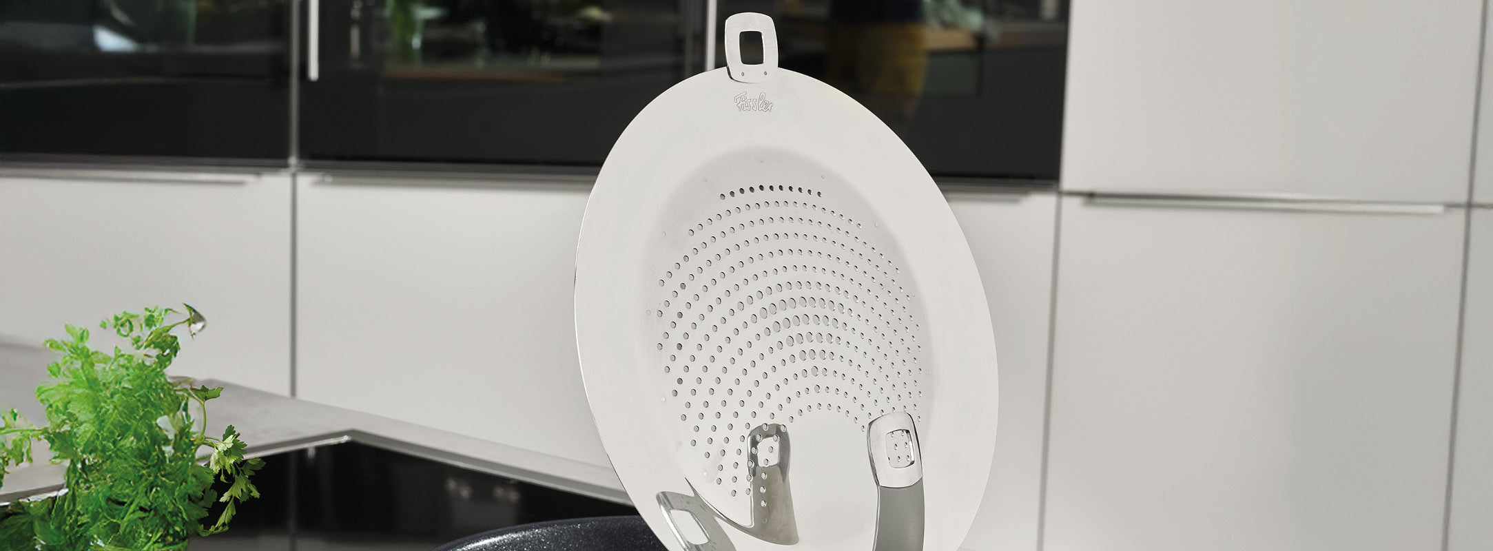 Ein weißer Silikon-Spritzschutz von Fissler ruht auf einer schwarzen Pfanne in einer modernen Küche mit Geräten aus Edelstahl und frischen Kräutern.