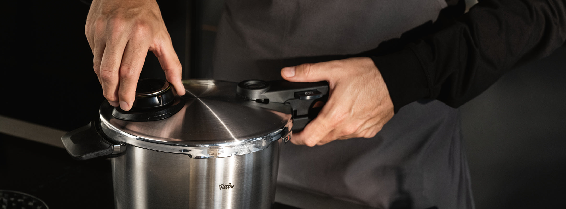 Eine Person in dunkler Schürze verschließt den Deckel eines Fissler Schnellkochtopfes aus Edelstahl und unterstreicht damit deutsche Präzision und Handwerkskunst.