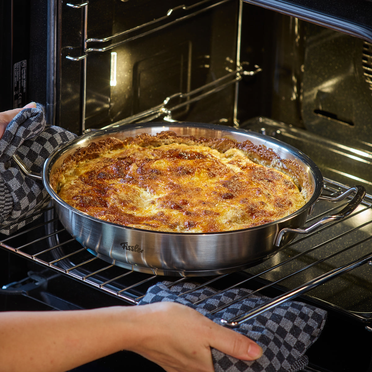 A person in checkered gloves slides a Fissler stainless steel pan with a golden, crispy casserole into the oven, showcasing German quality.