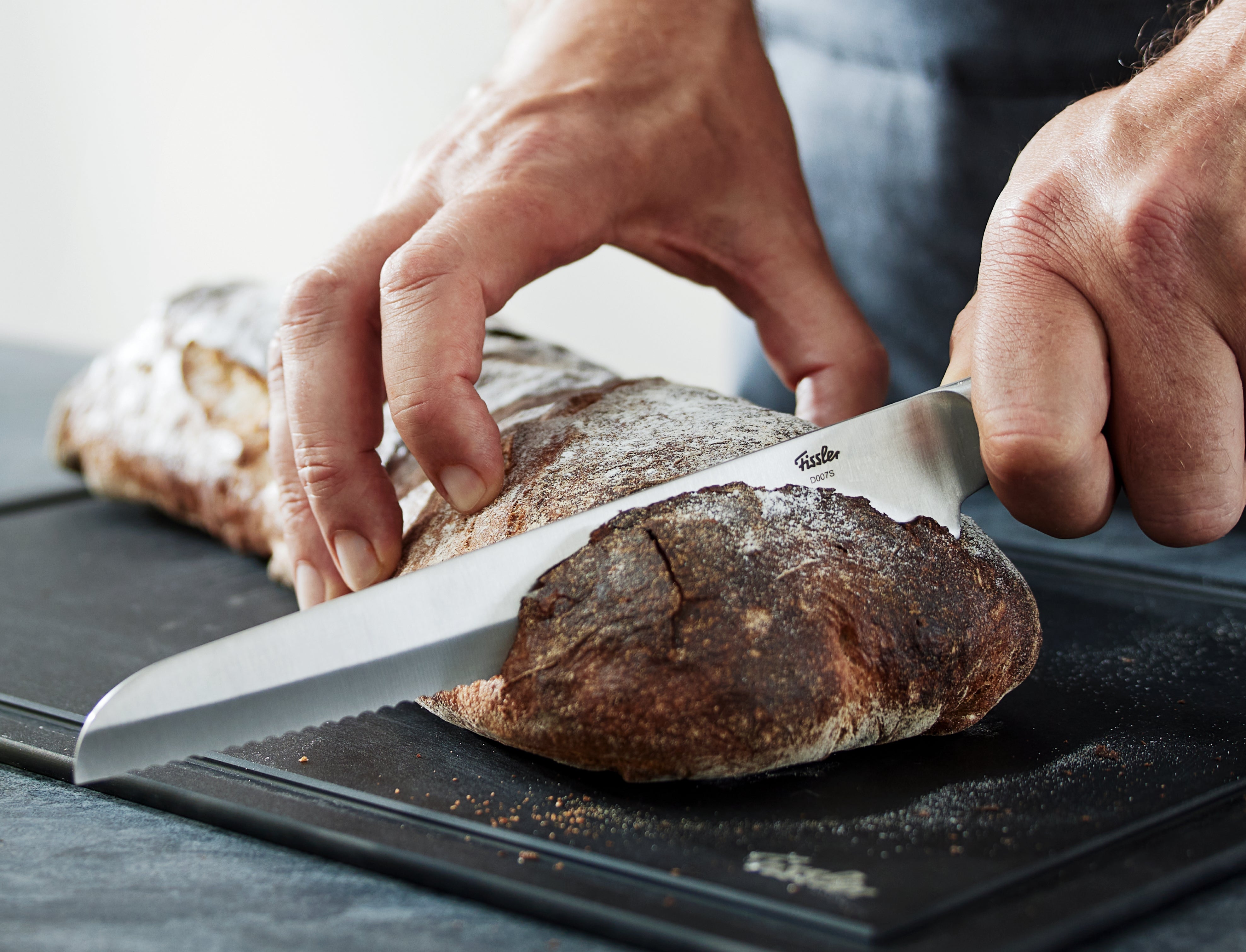 Eine Person schneidet rustikales Brot mit einem präzisen Wellenschliffmesser auf einem glatten Fissler-Schneidebrett; die Kruste ist strukturiert und mit Mehl bestäubt.